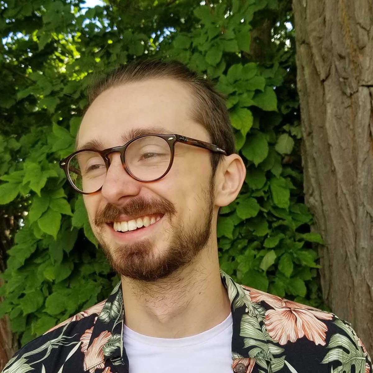 Photo of Carson Custer, a young white man with glasses and a bright floral shirt, smiling in front of greenery